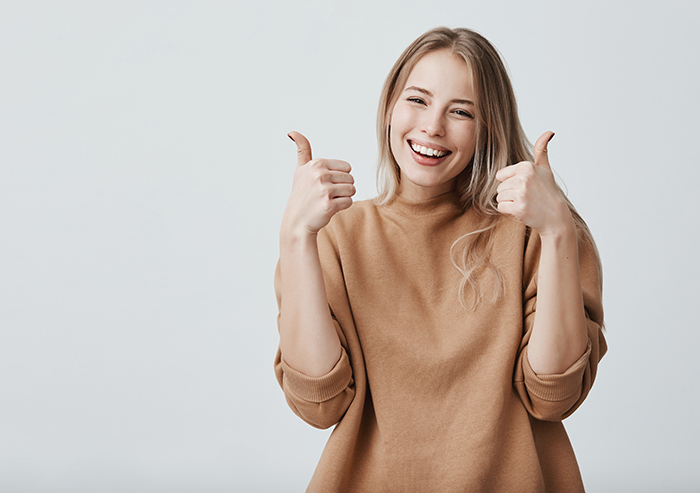 Portrait of fair-haired beautiful female student or customer with broad smile, looking at the camera with happy expression, showing thumbs-up with both hands, achieving study goals. Body language